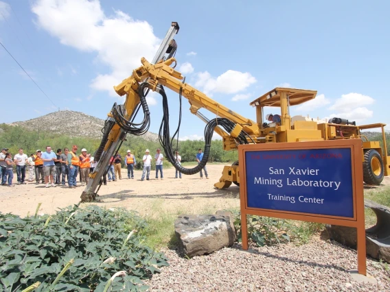 A group of people looking at an excavator behind a sign that says, "San Xavier Mining Laboratory."