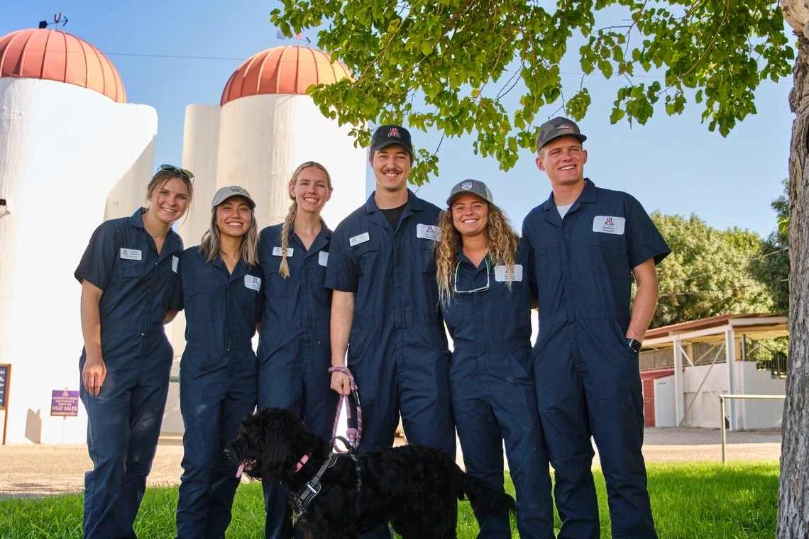 6 vet med students smile on farm with leashed black lab 