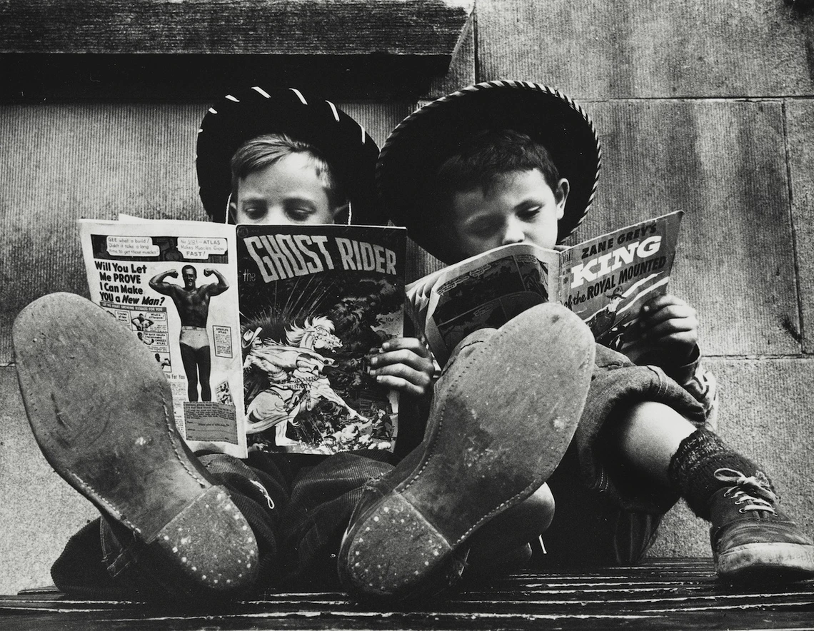 Two children wearing cowboy hats and boots, reading comics.